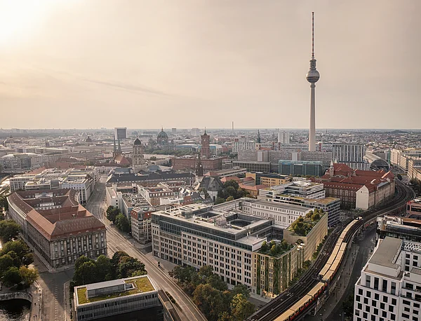 Luftaufnahme von Berlin mit Blick auf den Fernsehturm, umliegende historische Gebäude und moderne Bürohäuser bei leicht bewölktem Himmel.